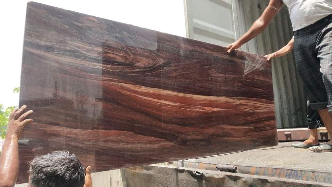 Polished teakwood granite slab with swirling wood-grain pattern being unloaded